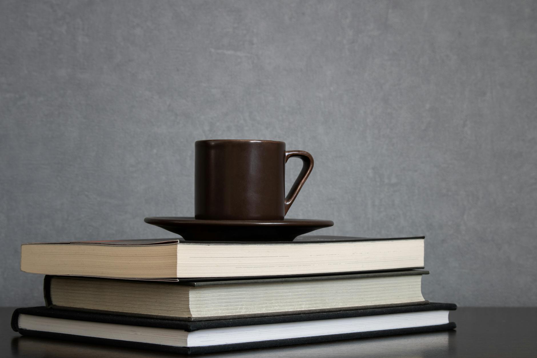 brown cup of coffee with saucer on top of pile of books to read placed off center of frame on a clean gray background empty space for text study reading or preparing for exams concept